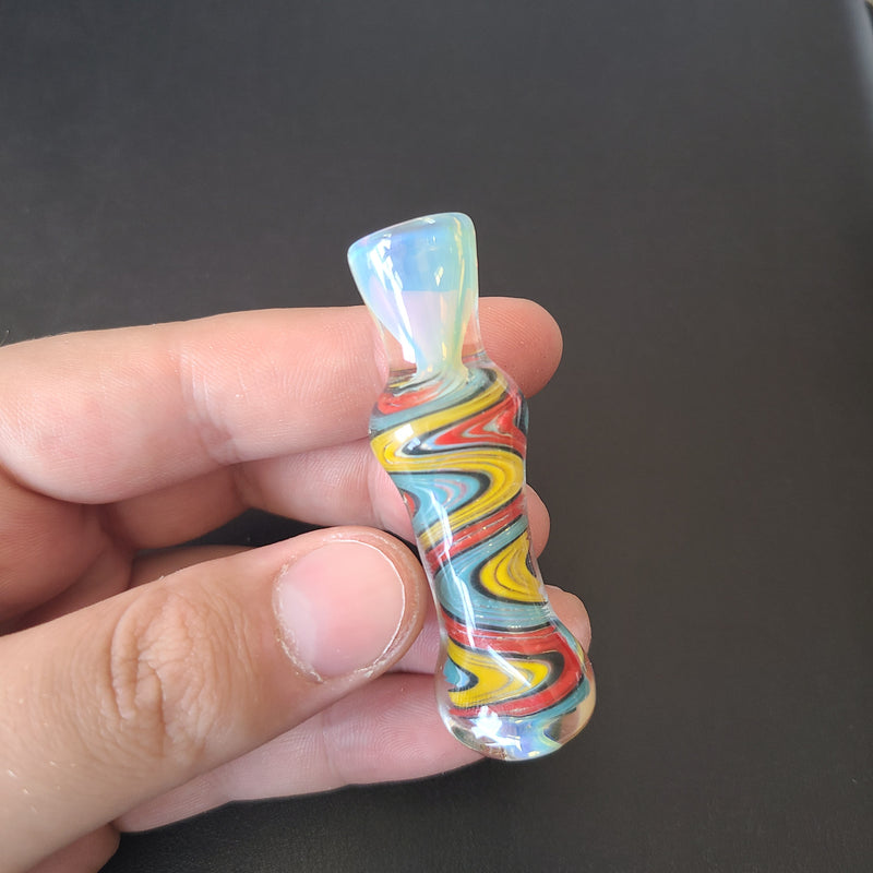 A small 3 inch Chillum Glass Pipe on a colorful shape and on a grey background between the fingers of a hand